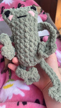 Handmade crochet frog toy held in a hand with a pink blanket in the background