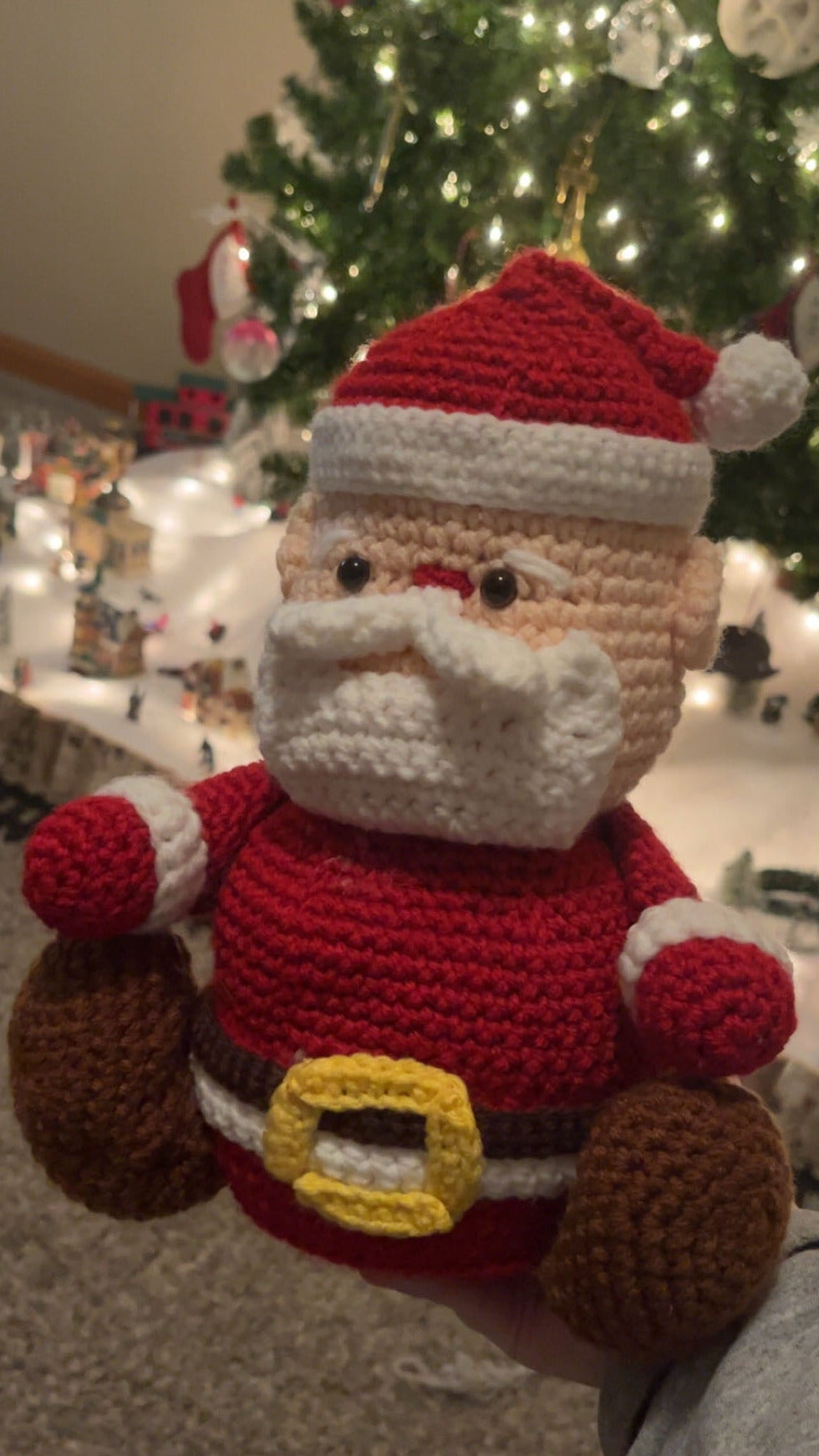 Crocheted Santa Claus figure held in front of a decorated Christmas tree.